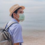 Man wearing a surgical mask and fedora backpacking on a serene beach, embracing the new normal.